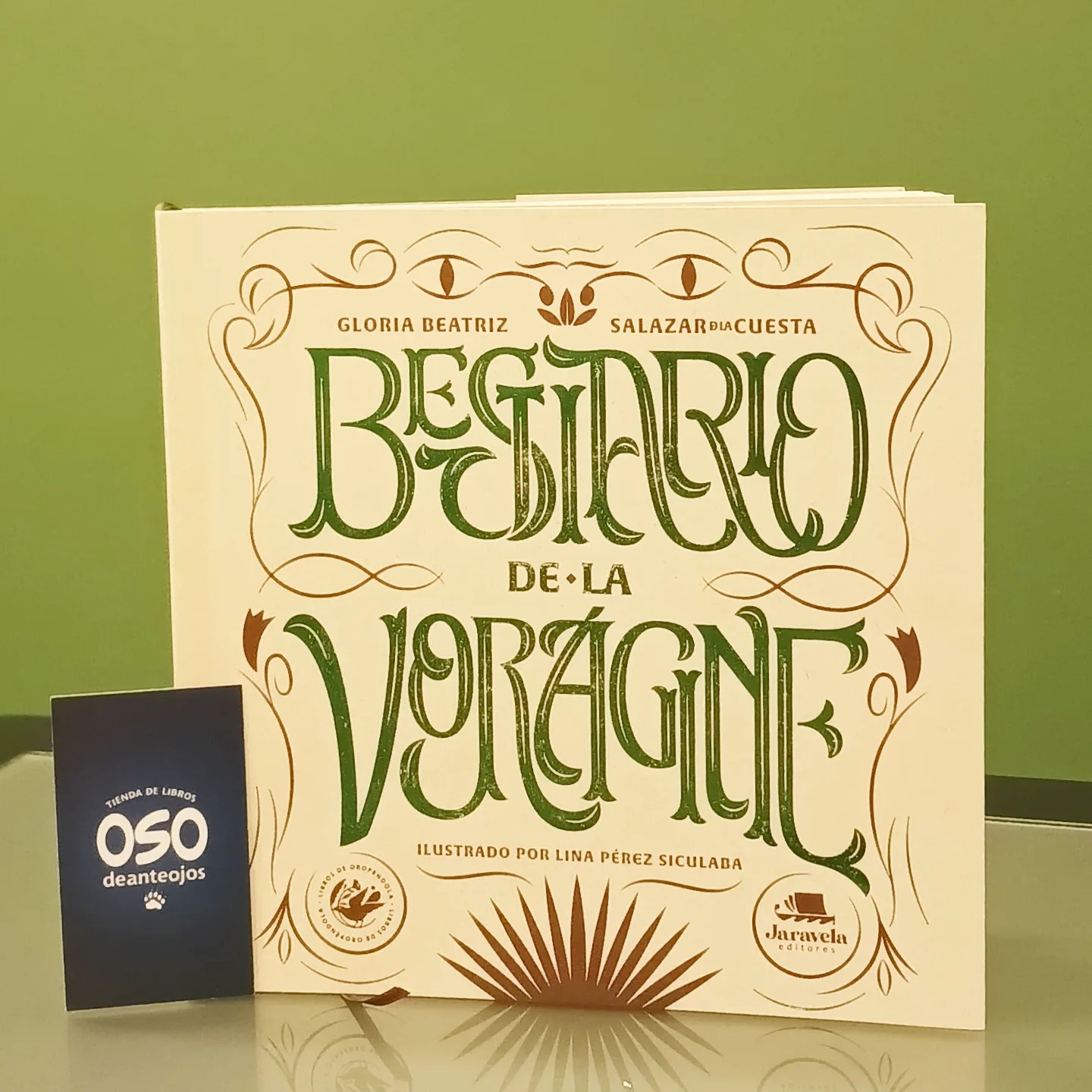 Fotografía del libro "Bestiario de La Vorágine" sobre una superficie clara. La portada es de color crema con tipografía elegante en tonos verdes y ocres, rodeada de ornamentos clásicos. Al lado izquierdo, se encuentra una pequeña tarjeta azul oscuro con el logotipo de la librería "Oso de anteojos".