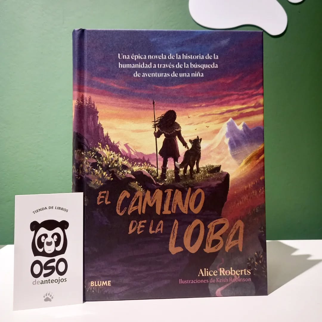Fotografía del libro "El camino de la Loba" de Alice Roberts, con ilustraciones de Keith Robinson, sobre una superficie blanca. La portada, de estilo artístico, muestra a una niña y su loba en un acantilado ante un paisaje montañoso y un atardecer vibrante. A la izquierda, una tarjeta blanca con el logotipo de la librería "Oso de anteojos".